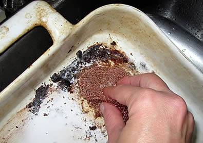 A hand is cleaning the oily corner of a tray, using a copper scrubber.